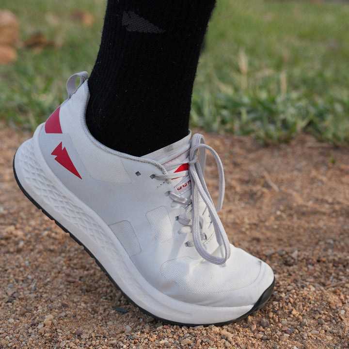 Close-up of a person showcasing the GORUCK Rough Runner shoes in Light Grey + Red, featuring a Gradient Density EVA midsole for comfort, paired with black socks on a gravel path. The shoes also boast a road-to-trail outsole.