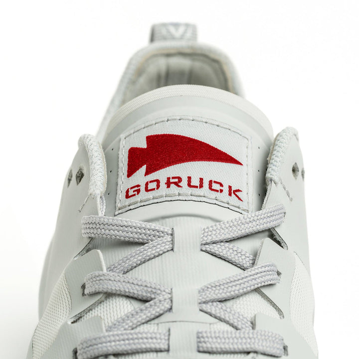 Close-up of a Rough Runner shoe in light grey, highlighted by gray laces and a red "GORUCK" logo on the tongue, featuring a road-to-trail outsole for versatile traction.