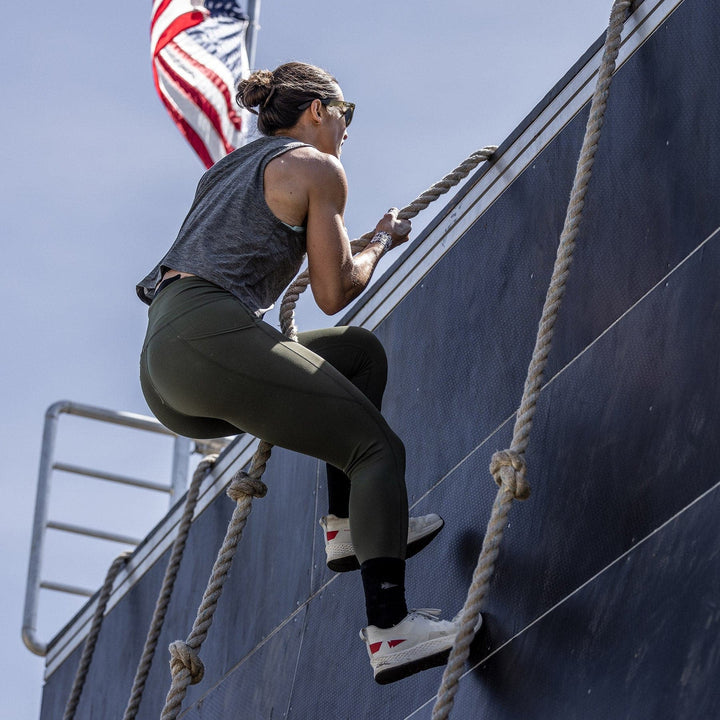 Wearing the Rough Runner - Light Grey + Red from GORUCK, which features a Gradient Density EVA midsole for ultimate support, a person climbs a rope against a wall with the U.S. flag waving proudly in the background.