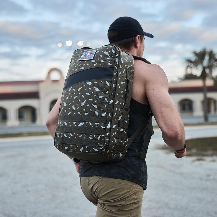 An individual with a patterned Rucker 4.0 from GORUCK is walking outdoors near a building and palm tree under a cloudy sky, embracing the spirit of rucking.