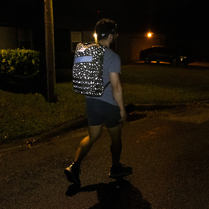 A person strolls at night on a wet street, casually dressed and sporting a patterned GORUCK Rucker 4.0 backpack.