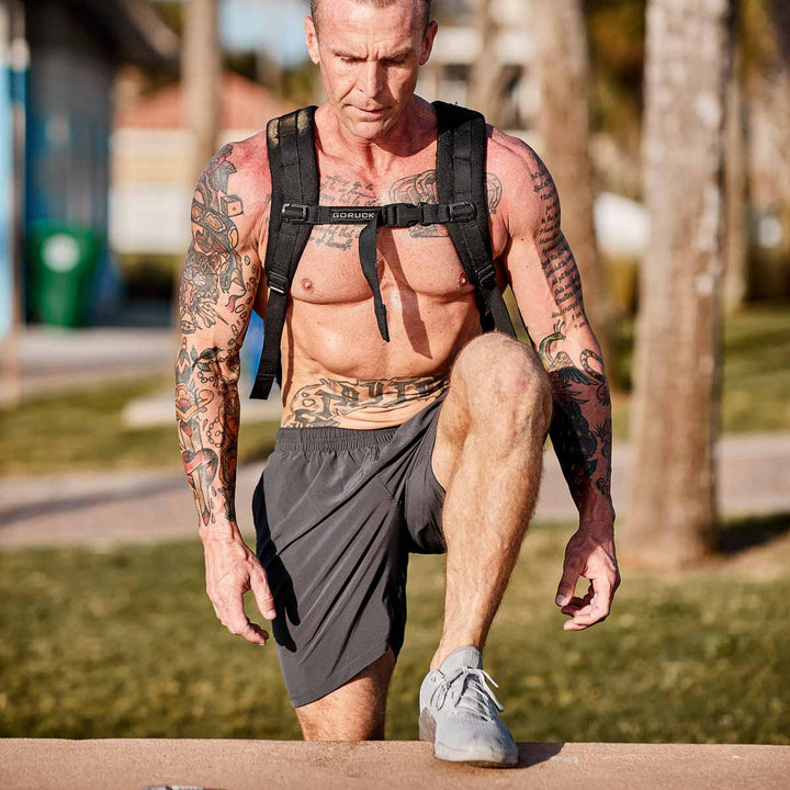 A muscular man with tattoos, sporting a GORUCK Ruck Plate Carrier 3.0 made from durable 1000D CORDURA and gray shorts, exercises outdoors. He steps onto a low wall amidst trees and grass on a sunny day, demonstrating the versatility of his carrier in this peaceful environment.