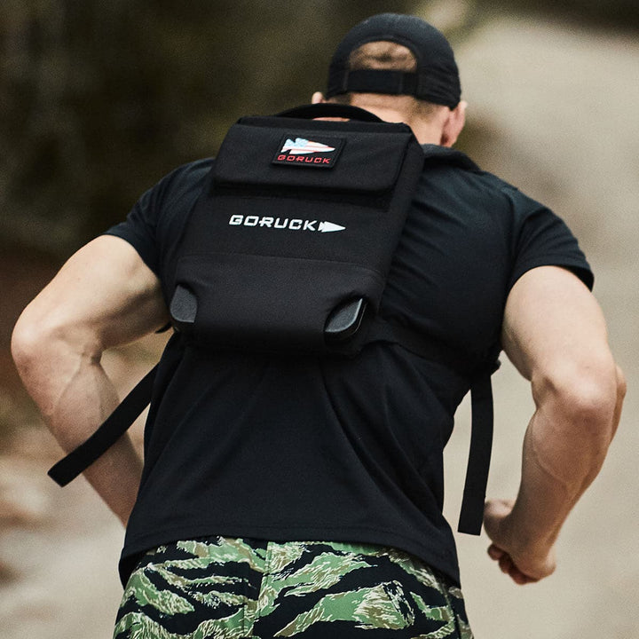 A person with a black Ruck Plate Carrier 3.0 by GORUCK, featuring ergonomic lumbar support, and wearing camouflage shorts is running on a path. The individual is seen from behind, sporting a cap on their head.