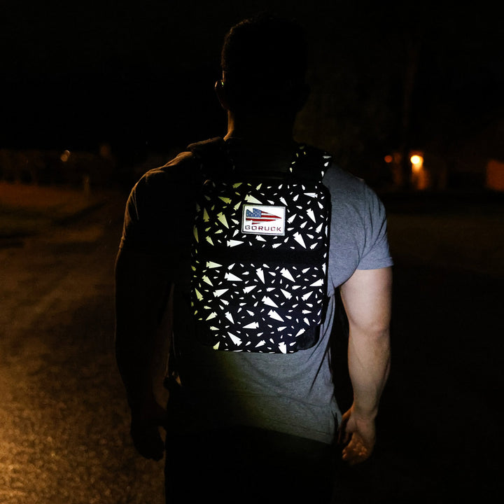 An individual walks through the night carrying their Ruck Plate Carrier 3.0 by GORUCK, adorned with a small American flag patch and equipped with a padded top handle for effortless portability.