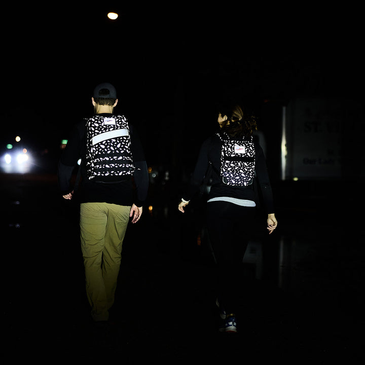 Two individuals wearing GORUCK Ruck Plate Carrier 3.0 packs are walking at night, illuminated by headlights against a dark background. These carriers provide ergonomic lumbar support and include a padded top handle, ensuring a comfortable and efficient midnight trek.