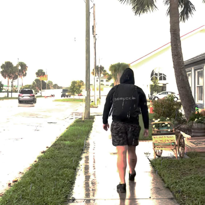 A person wearing a black hoodie and backpack with the GORUCK Ruck Rain Cover strides across a wet sidewalk dotted with puddles. They are moving away from the camera. To the right, there's a small cart adorned with flowers. Palm trees border the street on this cloudy day.