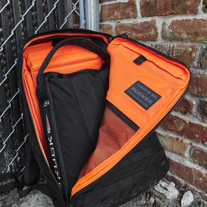 A Rucker 4.0 backpack in black with a vibrant orange interior is propped against a brick wall and metal fence. This GORUCK bag includes a mesh pocket and proudly features the Rucker® label, emphasizing its durable design ideal for rucking adventures.