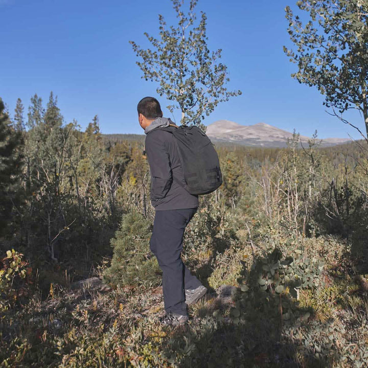 A person wearing a black jacket and GORUCK Men's Simple Pants - Midweight ToughDry® with a backpack stands on a rocky hill, surrounded by trees and foliage. In the background, there is a clear blue sky and a distant mountain peak.