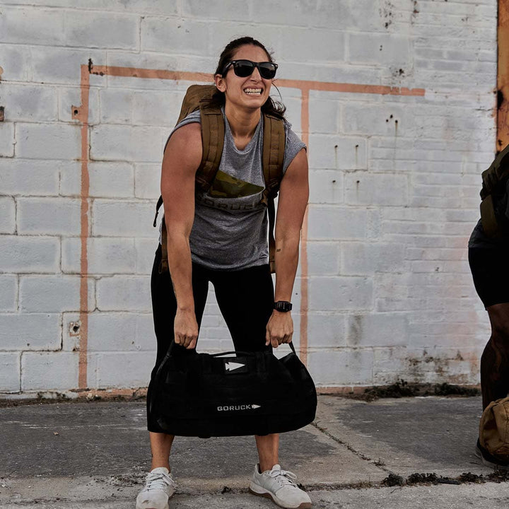 A woman smiles outdoors in a gray shirt, black leggings, and sunglasses as she lifts a Sandbag Kit by GORUCK with heavy-duty handles.