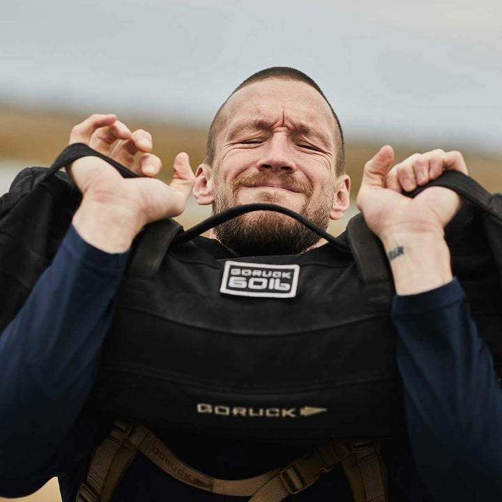 A person lifts a GORUCK Sandbag Kit (w/ Filler Bag) outdoors, grimacing with effort. The durable handles enhance grip, while the Scars Lifetime Guarantee assures lasting performance.