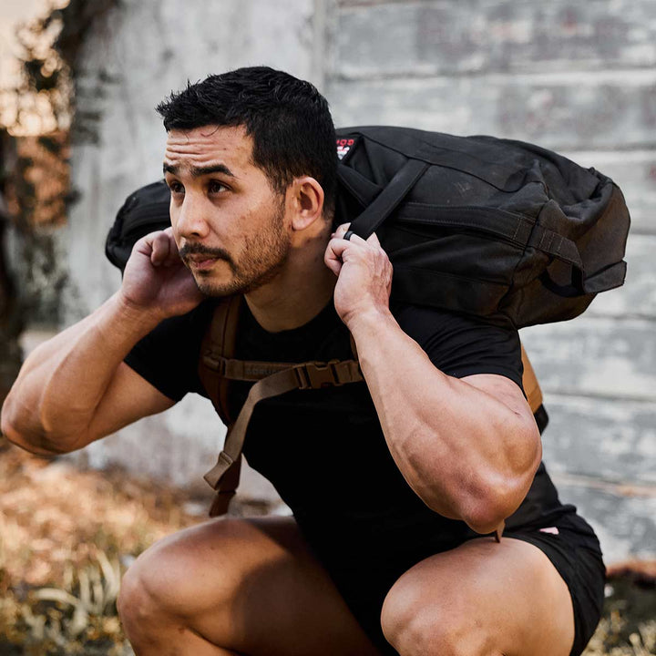 A man squats outdoors with a Sandbag Kit (w/ Filler Bag) from GORUCK featuring heavy-duty handles on his shoulders.