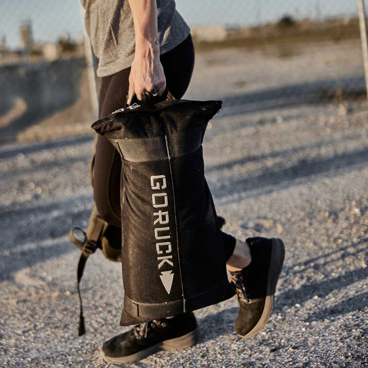 An individual with a GORUCK Simple Training Sandbag, protected by the Scars Lifetime Guarantee, is outside in sport attire, strolling along a gravel path.