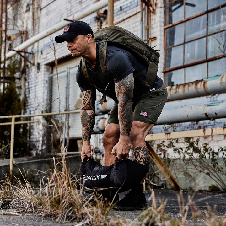 Amidst industrial buildings and overgrown plants, a man embodies the spirit of an outdoor home gym as he lifts GORUCK's Simple Training Sandbags, while wearing a backpack and cap.