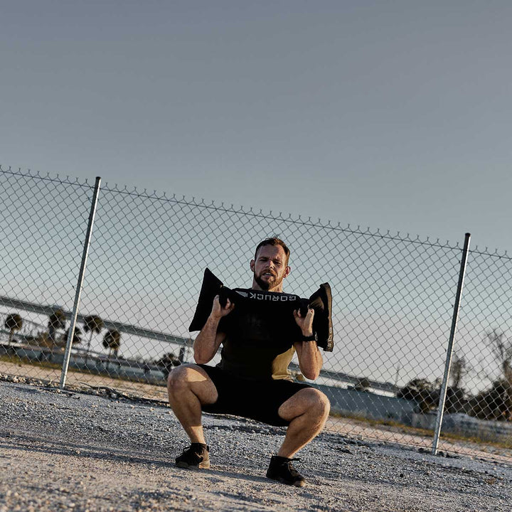 A man exercises outdoors by squatting with a GORUCK Simple Training Sandbag, dressed in fitness gear against a chain-link fence and under a clear sky. This innovative GORUCK product is ideal for your home gym, offering durability and versatility for all types of workouts.