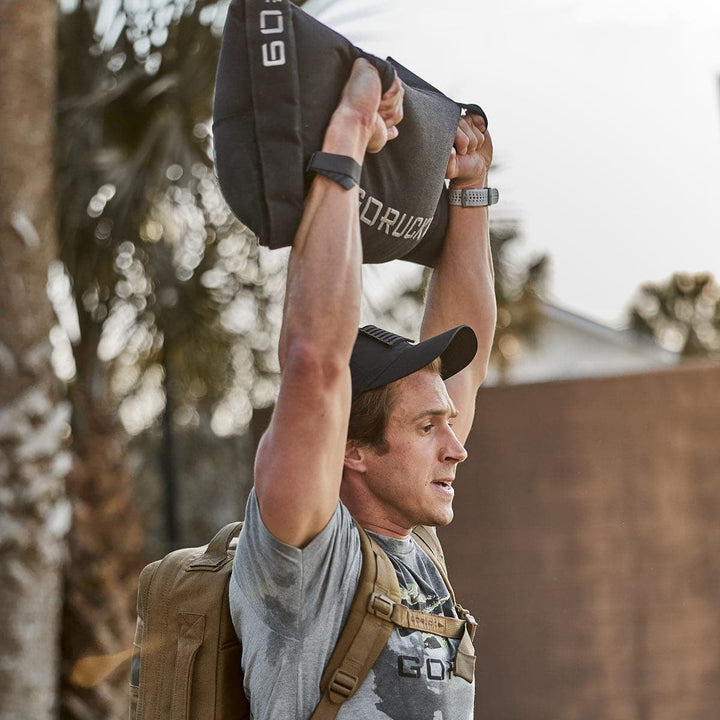 A man, dedicated to his outdoor training routine, effortlessly lifts a Simple Training Sandbag by GORUCK overhead. Wearing a hat and backpack from the Scars Lifetime Guarantee collection, he stands amidst the trees, fully embracing nature as his personal gym.