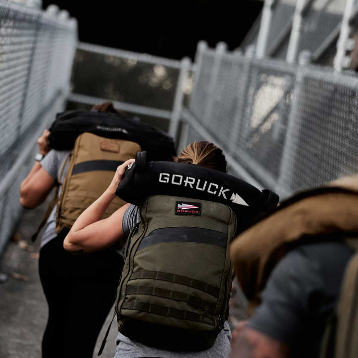 Individuals carrying GORUCK backpacks and Simple Training Sandbags while walking along a fenced pathway.