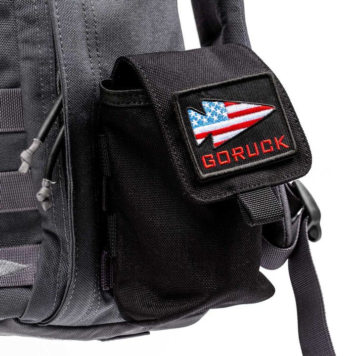 The Simple Side Pocket in black, featuring the GORUCK logo with an American flag design, is attached to a gray backpack with internal organization. It is securely fastened using VELCRO®.