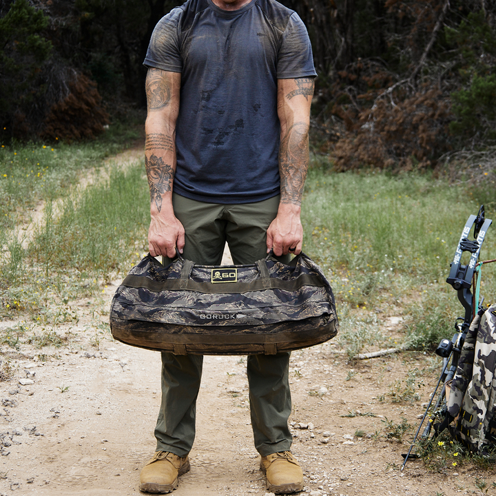 A person in a dark shirt and green Men’s Simple Pants by GORUCK stands on a dirt path enveloped by lush foliage, holding an abrasion-resistant, camouflage duffel bag. Tattoos are visible on their arms, with GORUCK's hiking gear made from Lightweight ToughDry® fabric nearby.