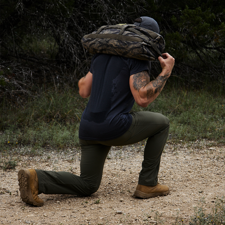 A person dressed in a black t-shirt, GORUCK’s Men’s Simple Pants – Lightweight ToughDry® in green, and tan boots kneels on one knee outdoors with a camouflage-patterned bag on their shoulders. The ToughDry fabric of their pants provides abrasion resistance against the grassy and tree-filled backdrop.