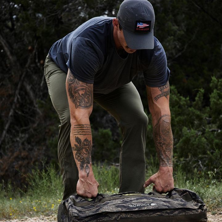 A person wearing a cap and dark t-shirt bends down to lift a camouflage-patterned bag outdoors, standing on a gravel surface and surrounded by trees. Tattoos decorate their arms and neck. The bag is made from GORUCK's abrasion-resistant ToughDry® fabric, ideal for rugged adventures.