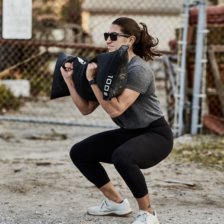 Wearing sunglasses, a person is exercising outdoors with the 100-pound Simple Training Sandbags by GORUCK, which features padded handles. They are squatting with a chain-link fence and some outdoor equipment visible in the background.