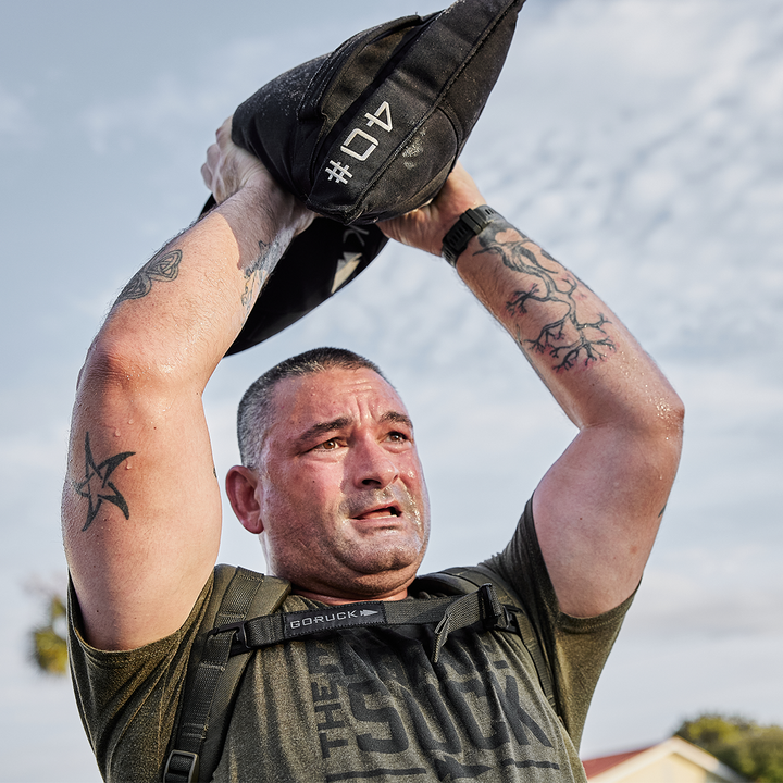 A person wearing a green T-shirt lifts a GORUCK Simple Training Sandbag overhead. Tattoos adorn both arms as they focus with intensity under the cloudy sky, beads of sweat marking their dedication to home gym workouts.