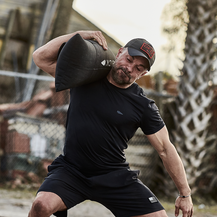 A man in GORUCK athletic wear and a cap lifts a Simple Training Sandbag with padded handles onto his shoulder outdoors. He appears focused and determined, set against a fenced background with a palm tree nearby.