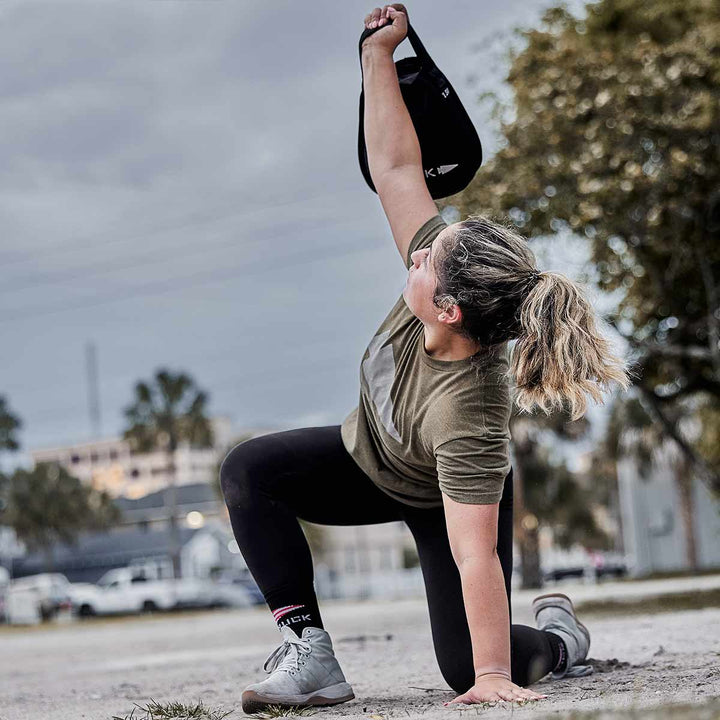 A person in workout gear kneels on one knee, effortlessly lifting a Sand Kettlebell by GORUCK overhead outdoors, showcasing its endurance design.