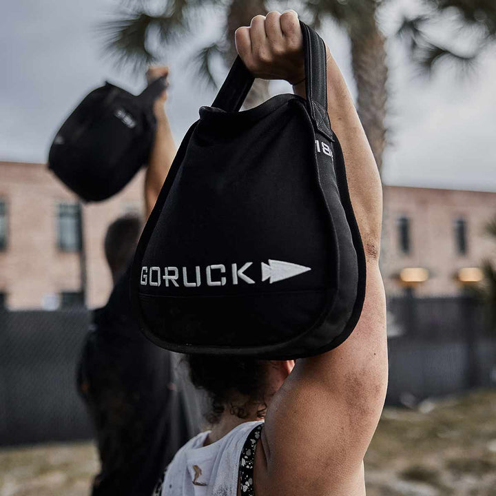 In a remarkable demonstration of strength, a person effortlessly lifts a GORUCK Sand Kettlebell outdoors, set against palm trees and a towering building that create a picturesque scene highlighting the versatility of this portable kettlebell.