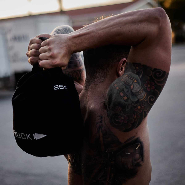 A man with arm tattoos skillfully lifts a 26-pound GORUCK Sand Kettlebell over his shoulders outdoors, highlighting its durable design.