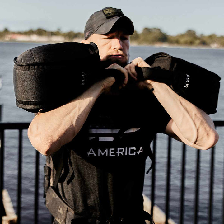 By the tranquil lake, an individual lifts a GORUCK Sand Kettlebell, pushing their endurance to new heights. Dressed in a black shirt with "AMERICA" emblazoned across it and a cap, they embrace the challenge of outdoor fitness with determination.