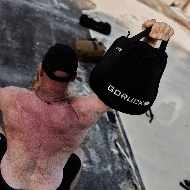 A man showcases the durability and versatility of GORUCK's Sand Kettlebells as he lifts a black weighted bag on a concrete surface outdoors.