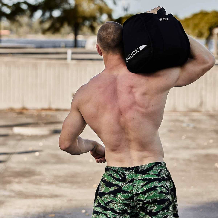 A shirtless man outdoors confidently shoulders a GORUCK Sand Kettlebell - 53LB (24KG), showcasing his strength in green patterned shorts.