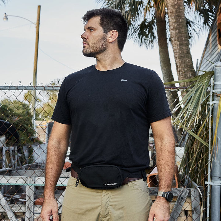 A man in a black shirt and khaki pants stands outside near a chain-link fence and palm trees, casually sporting the GORUCK Belt Bag made with Ripstop ROBIC®, slung across his shoulder.