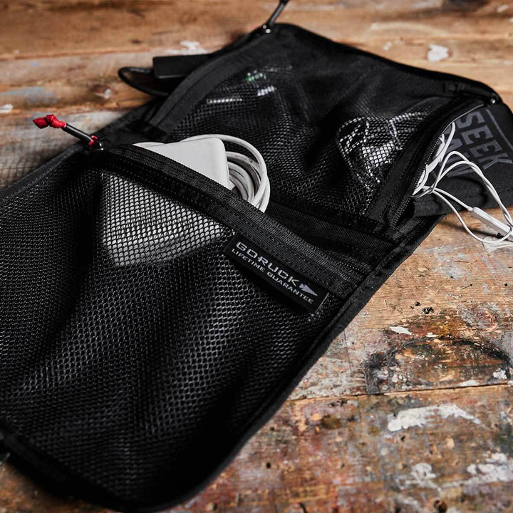 A black GORUCK Wire Dopp, crafted from durable CORDURA, rests on a wooden surface, neatly storing electronic accessories such as a white mouse and cables. The brand's label is prominently displayed, making it an ideal choice for organizing your travel essentials.