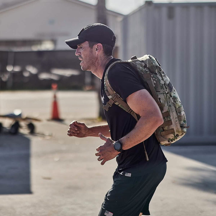 A man wearing a black Performance TAC Hat - TOUGHDRY from GORUCK and a matching shirt jogs outdoors, carrying a camouflage backpack made with sweat-wicking material to keep him cool. He appears focused, with weights on the ground nearby. A traffic cone and a building can be seen in the background.