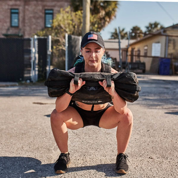 A person squats outdoors while lifting a heavy sandbag, wearing GORUCK's Performance TAC Hat - TOUGHDRY, a sweat-wicking cap with an American flag patch, along with a sports bra, shorts, and sneakers. In the background are a brick building and fence illuminated by bright sunlight.