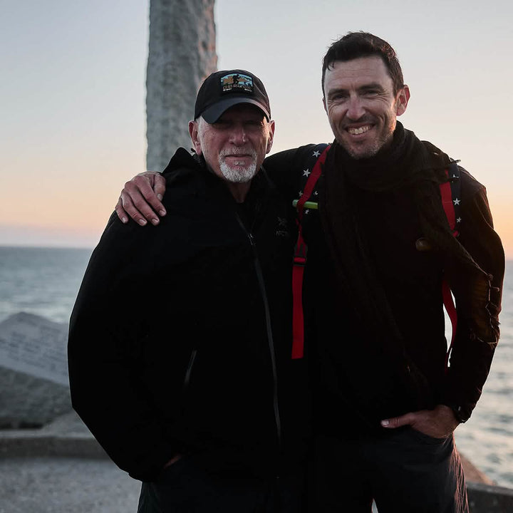 Two men stand side by side, grinning by the seaside. One is donning a GORUCK Performance TAC Hat in black along with a matching military-style jacket, while the other showcases a sweat-wicking black scarf paired with a backpack. The ToughDry® material ensures they remain cozy against the ocean breeze, set against a stone monument and clear sky backdrop.