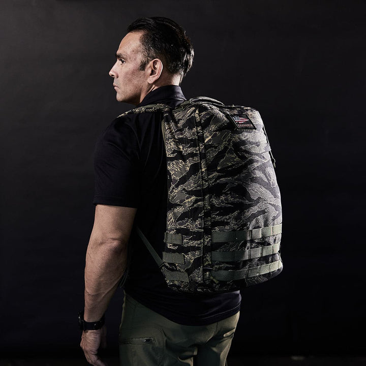 A person in a black shirt and green pants wears a GR1 USA rucksack from GORUCK crafted from 210D CORDURA, glancing to the side against a dark background.