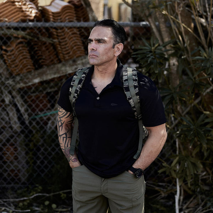 A person with a GR1 USA rucksack by GORUCK stands outdoors, wearing a black shirt and green pants, beside a chain-link fence and trees.