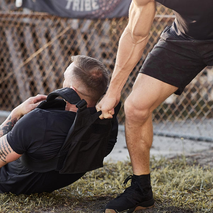 A person pulls another across the ground using a GORUCK Training Weight Vest outdoors near a chain-link fence.