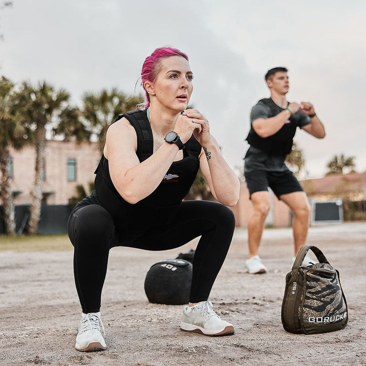 Individuals dressed in gym attire and the GORUCK Training Weight Vest 2.0 practice squats outdoors, surrounded by exercise equipment.