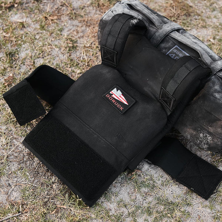 The GORUCK Training Weight Vest 2.0, featuring the "GORUCK" logo, rests on a grassy surface and is crafted to hold Ruck Plates for improved training performance.