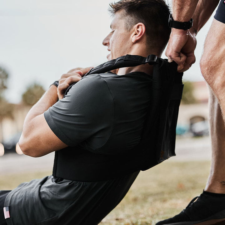 A man in workout gear is doing resistance training outdoors, wearing the Training Weight Vest 2.0 from GORUCK, while someone assists him.