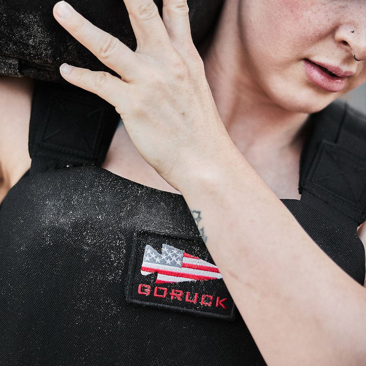 A determined individual carrying a heavy load, sporting a GORUCK Training Weight Vest 2.0 in black with an American flag patch and fitted with Ruck Plates for added resistance.