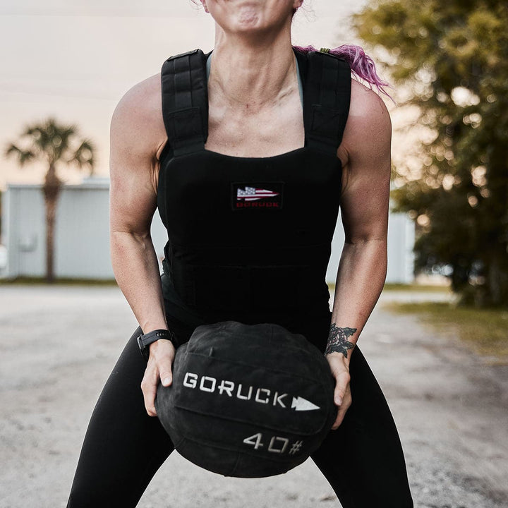 An individual sporting the Training Weight Vest 2.0 from GORUCK hoists a 40-pound ball outdoors, demonstrating their strength and commitment.