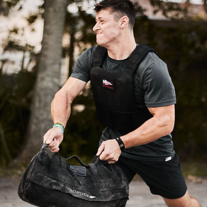 A man wearing the GORUCK Training Weight Vest 2.0 lifts a heavy sandbag outdoors, surrounded by trees in the background.