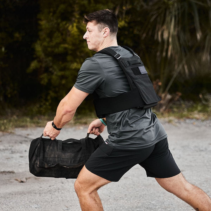 A man in a GORUCK Training Weight Vest 2.0 sprints with determination, carrying a black duffel bag outdoors.