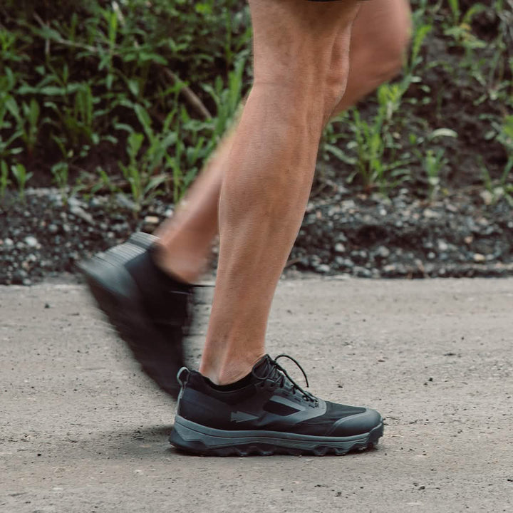 A person runs along a dirt path wearing GORUCK Mackall shoes in Black + Forged Iron, featuring a triple compound outsole for enhanced grip and durability.