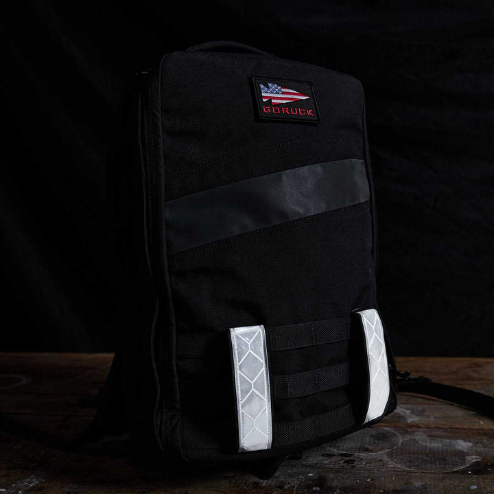 A black GORUCK backpack equipped with reflective Ruck Bands for enhanced nighttime visibility and a patch showcasing the American flag and brand logo on a dark backdrop, complete with MOLLE webbing.